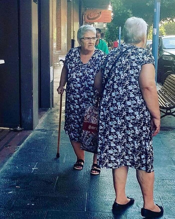 Two elderly women in identical floral dresses creating a weirdly hilarious visual illusion on a city sidewalk.