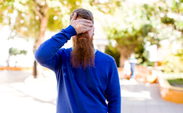 A bearded man in a blue sweater covering his eyes outdoors representing wildest dating experiences and disbelief.