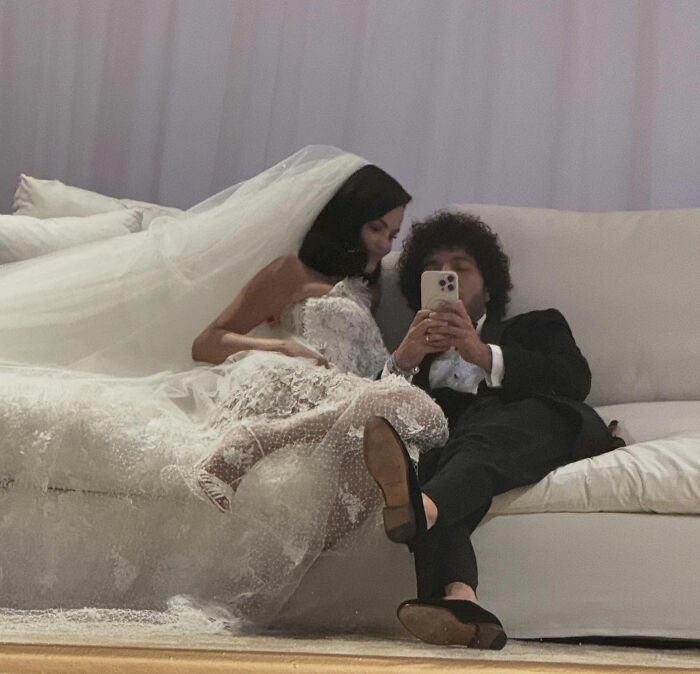 Benny Blanco and Selena Gomez in wedding attire relaxing on a couch, with Benny holding a phone for a selfie.