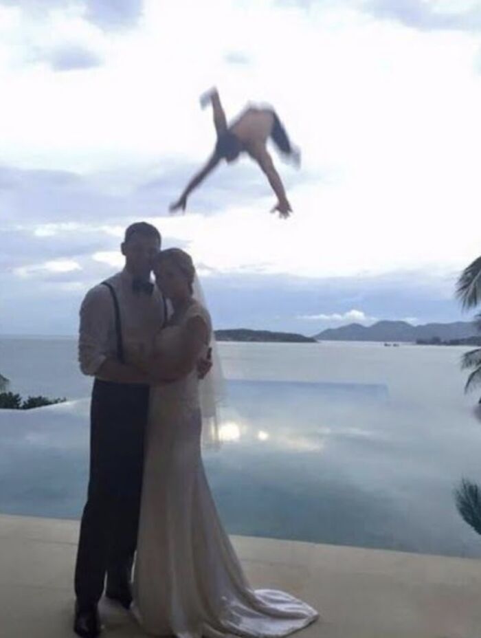 Couple posing at wedding by a pool with a person hilariously mid-flip in the background, weirdly funny image.