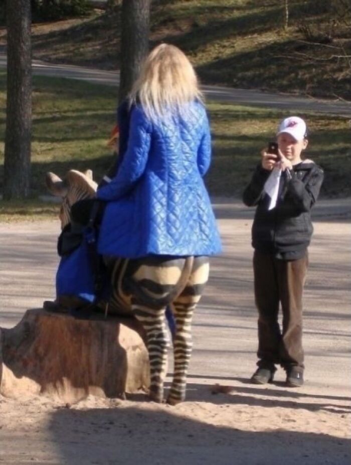 Woman in a blue coat standing behind a zebra statue while a boy takes a photo, creating a weirdly hilarious optical illusion.