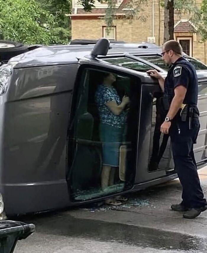 Police officer talking to a woman standing inside a flipped car in a weirdly hilarious images scene on a street.