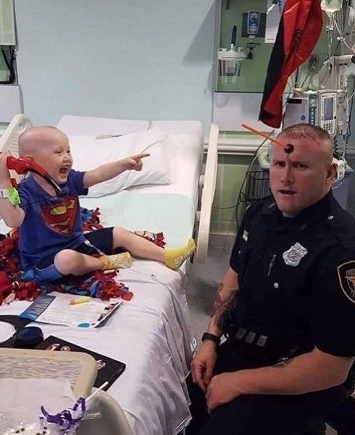 Child in hospital bed wearing Superman shirt laughs pointing at police officer with toy arrow stuck to his forehead in weirdly hilarious image.