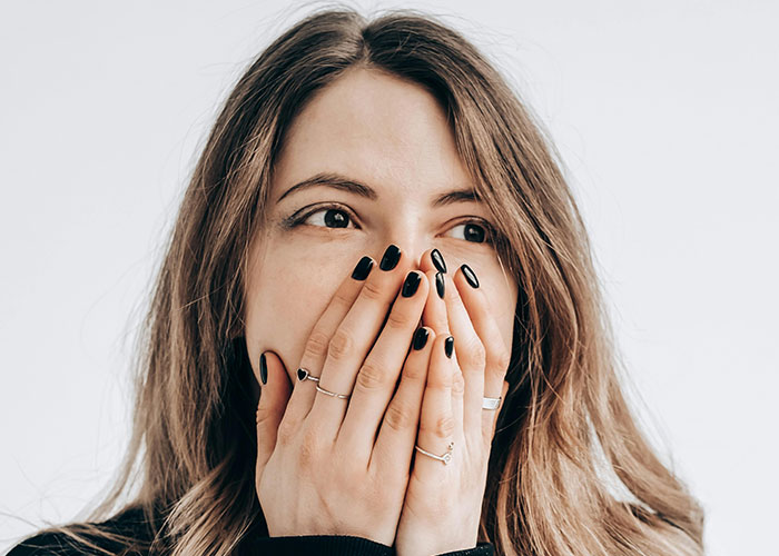 Woman with black nail polish and rings covering her mouth, surprised by a travel encounter with a kind stranger on a plane.