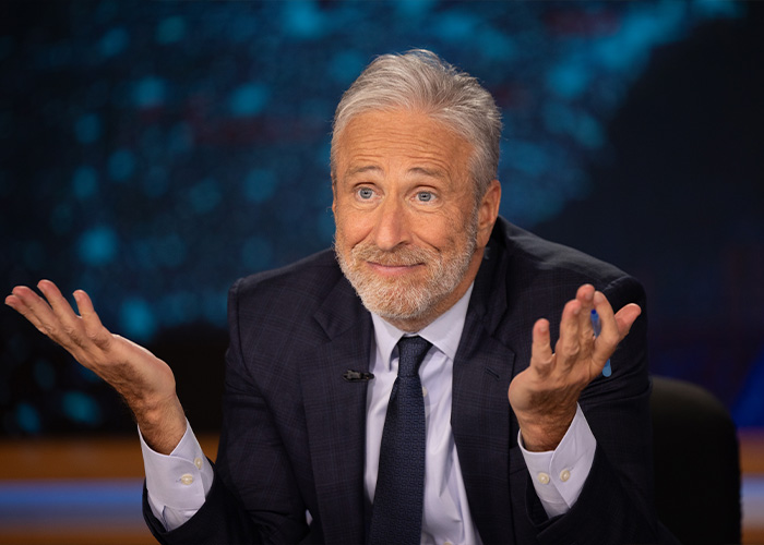 Jon Stewart in a suit shrugging with a slight smile during a televised interview about Epstein files mention.
