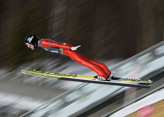 Ski jumper in red suit soaring through the air during Winter Olympics, linked to scandal involving acid male enhancement.