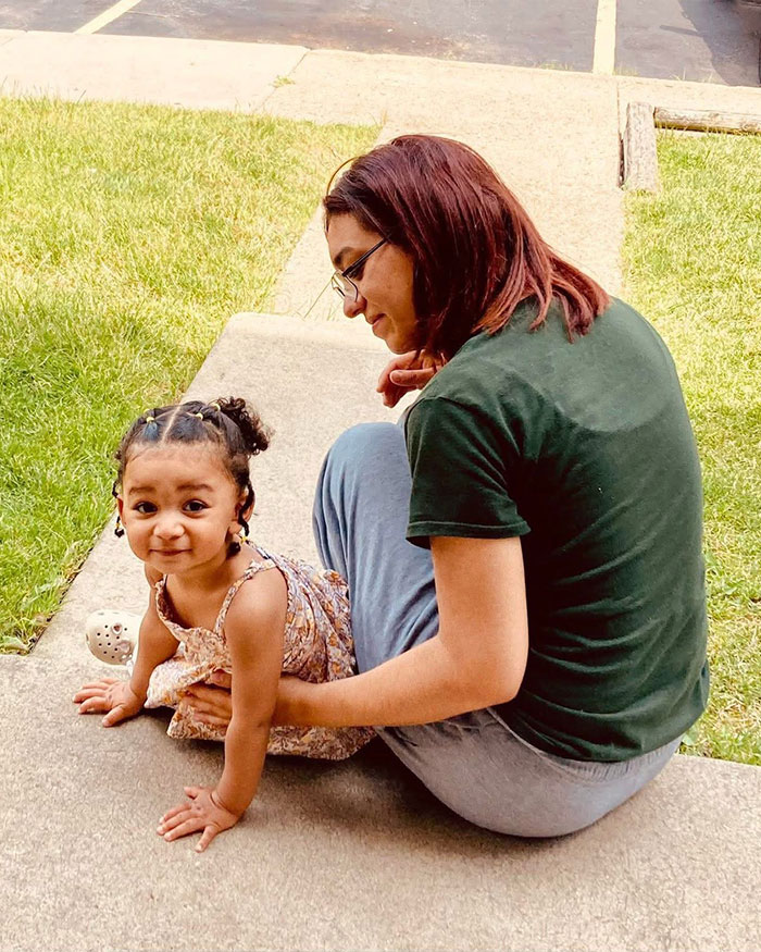 Missing woman 24 sitting outdoors with a young child on a concrete path near grass in casual clothing.