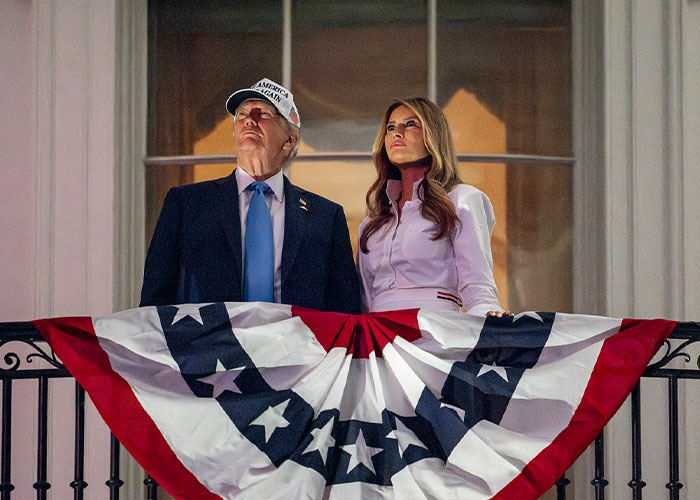 Donald Trump and Melania Trump stand on a balcony draped with patriotic bunting in a nighttime setting.