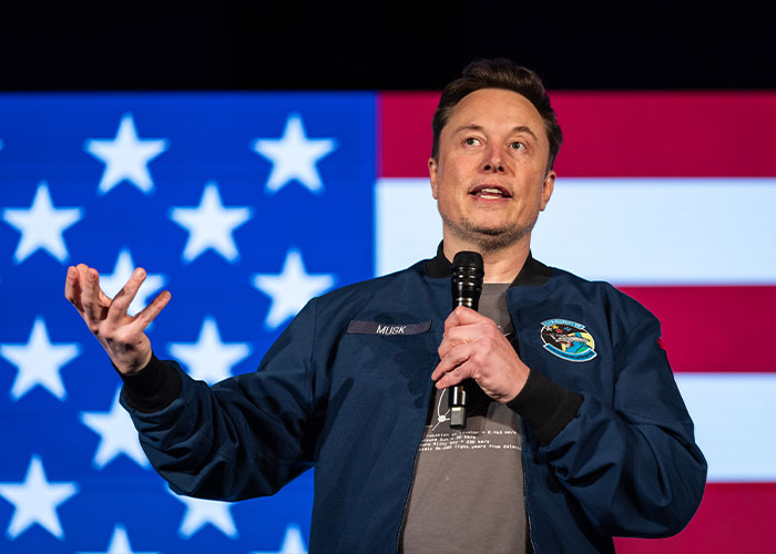 Elon Musk speaking on stage with an American flag background, sparking theories about something really bad coming.