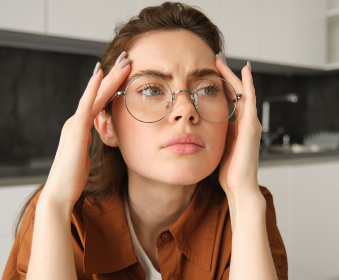 Young woman wearing glasses looking stressed and upset while sitting in a modern kitchen indoors.
