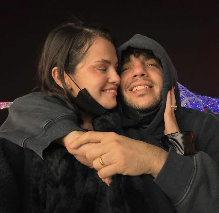 Benny Blanco and Selena Gomez sharing an affectionate moment outdoors at night with holiday lights in the background.