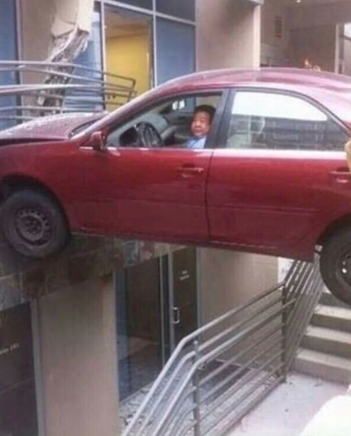 Man sitting in a red car oddly hanging over a stairwell railing, a weirdly hilarious image to improve your feed and mood.