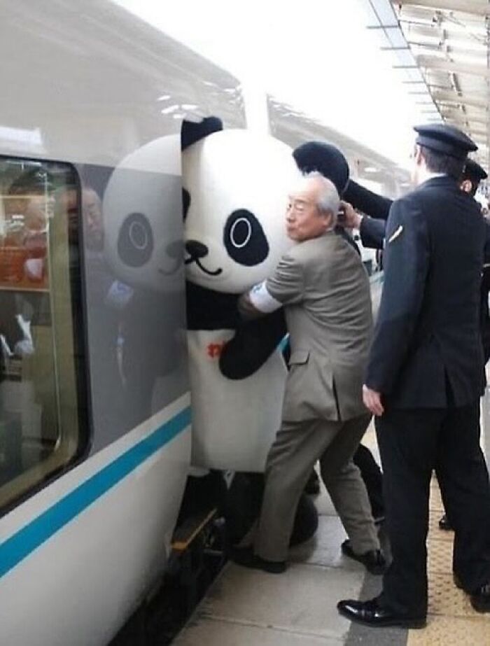 Man in suit hugging giant panda costume squeezed into train, a weirdly hilarious image at a busy platform.