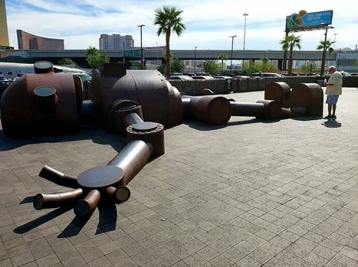 Giant quirky roadside attraction sculpture resembling a robot lying on pavement with palm trees and buildings nearby
