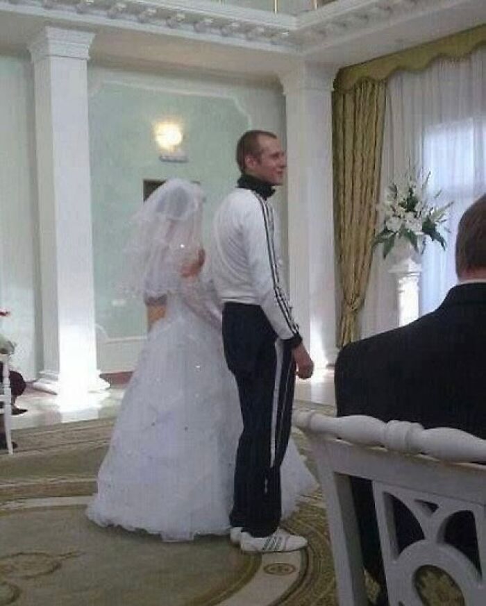 Bride in wedding dress standing next to groom wearing casual tracksuit and sneakers in a formal setting, weirdly hilarious image.