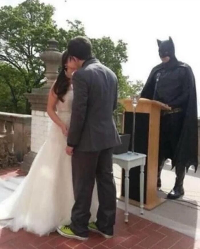 Couple kissing at wedding with groom wearing sneakers and person dressed as Batman officiating, weirdly hilarious image.