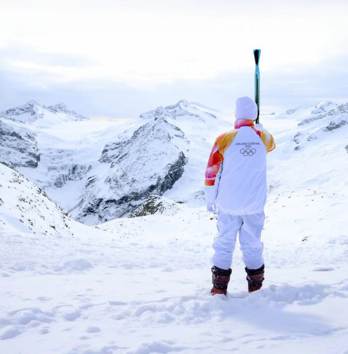 Winter Olympics athlete in uniform holding torch, standing on snowy mountain landscape during the event.