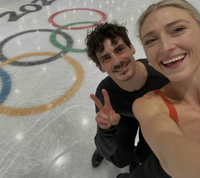 Olympic ice skater and partner smiling on the rink near Olympic rings after incredible save from wardrobe malfunction.