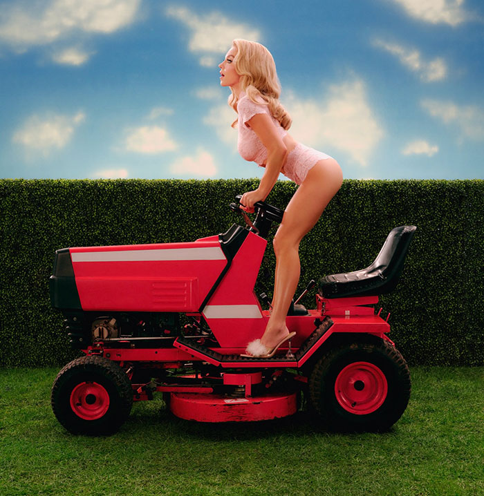Sydney Sweeney wearing pink lingerie posing on a red lawn mower against a green hedge and cloudy sky background.