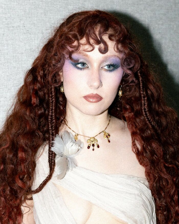 Chappell Roan with curly red hair and bold makeup, wearing a white dress and gold jewelry in an indoor setting.
