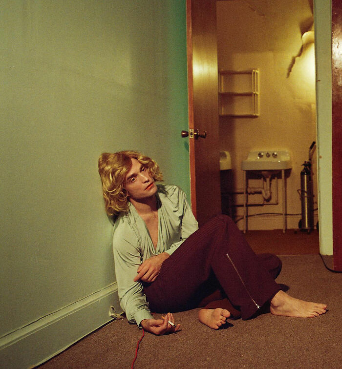 Robert Pattinson in a gender-bending photoshoot, sitting barefoot on the floor in retro clothing with a contemplative expression.