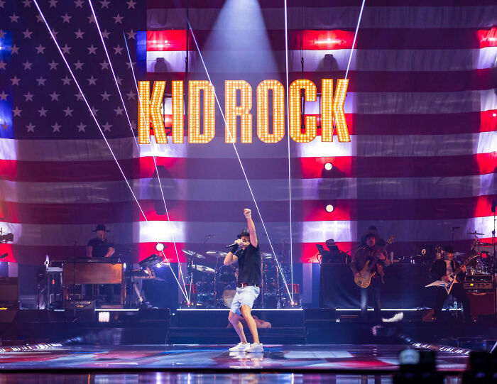 Kid Rock performing on stage with large illuminated name and American flag backdrop during alternative halftime show.