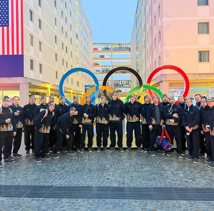 Olympic hockey rivals posing together in matching jackets in front of large Olympic rings sculpture outdoors.