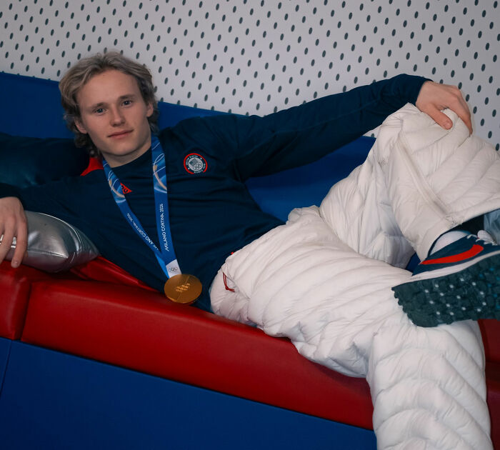 Ilia Malinin wearing a gold medal and casual sportswear, reclining on a red and blue couch in a relaxed pose.