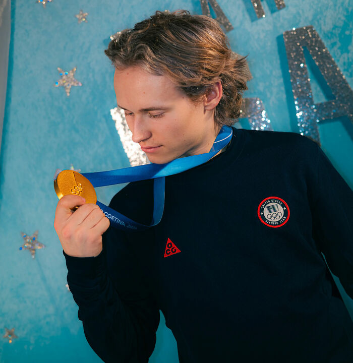 Ilia Malinin holding a gold Olympic medal, wearing Team USA apparel, with a blue starry background.