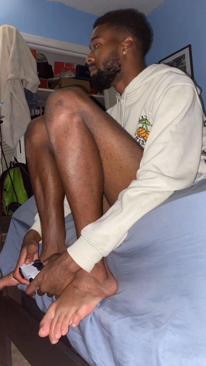 Man sitting awkwardly on bed with knees tucked to chest playing video games, showcasing funny men’s awkward body language.