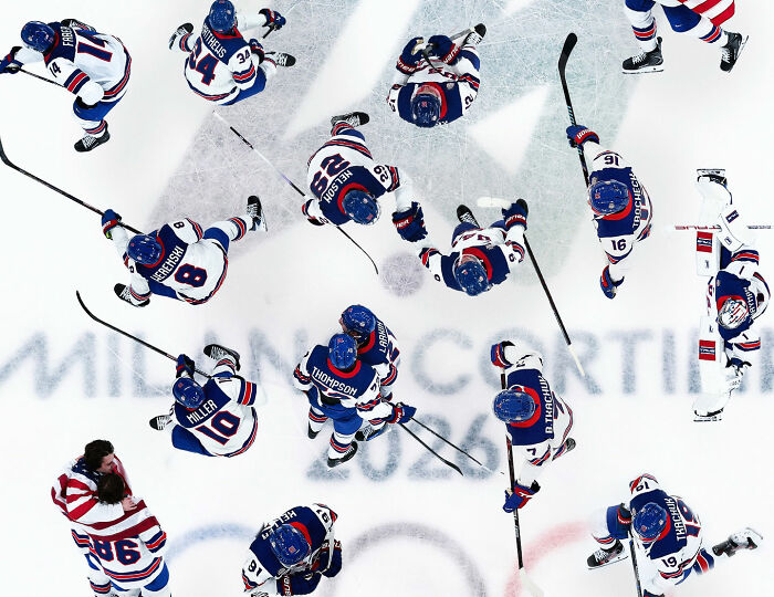 Team USA men's hockey players in blue jerseys huddle on ice with sticks during a game, seen from above. Team USA men's hockey players in blue jerseys huddle on ice with sticks during a game, seen from above.