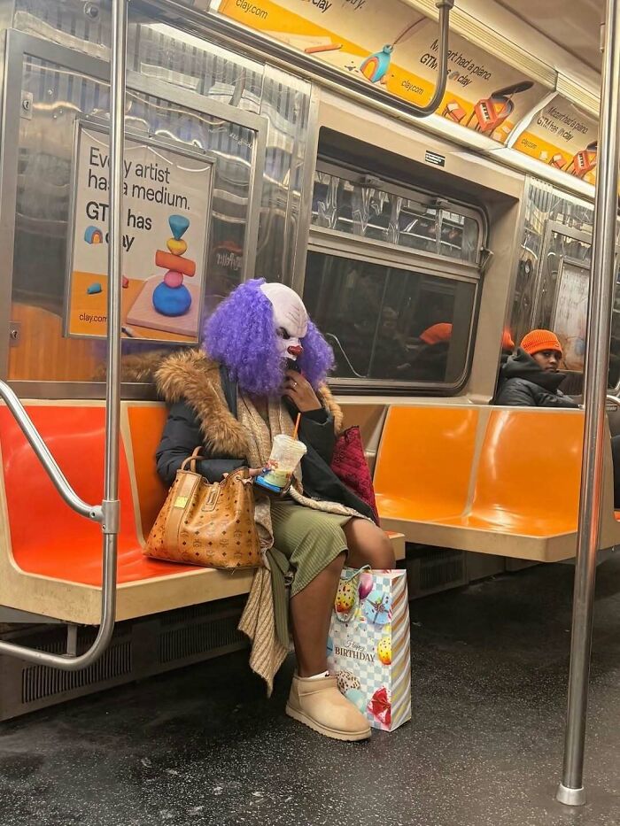 Person wearing a purple wig and clown mask sitting on a NYC subway bench with a birthday gift bag and drink.