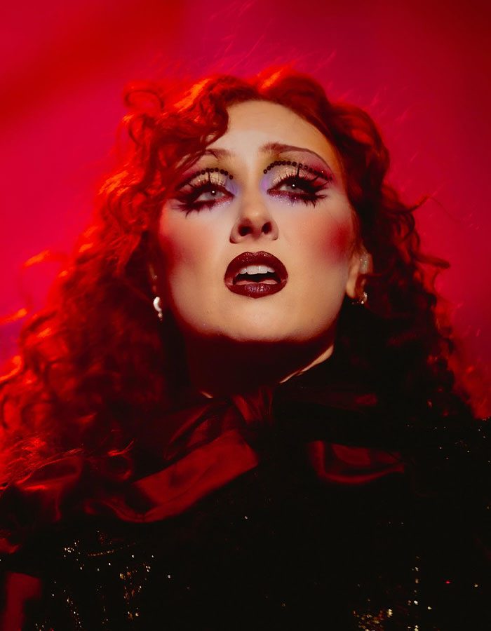 Chappell Roan with bold makeup and red curls under dramatic red lighting, body language expert analyzes Grammys 2026 appearance.
