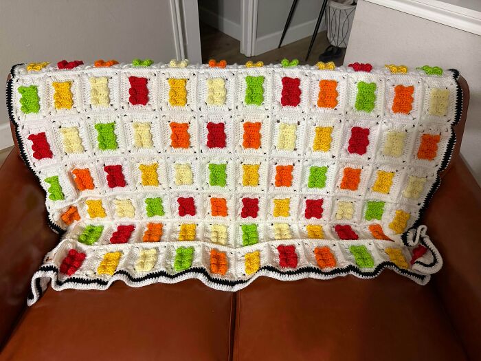 A white crocheted blanket with colorful gummy bear shapes, showcasing an awesome crocheted item on a brown sofa.