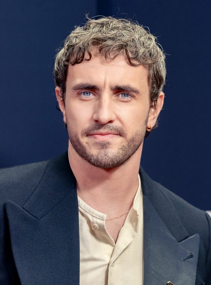 Paul Mescal with curly hair and beard, wearing a black jacket and light shirt, posing against a dark background.