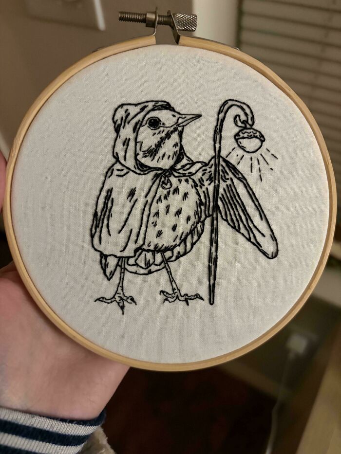 A charming embroidery creation of a bird in a cloak holding a lamppost, a whimsical embroidery design.