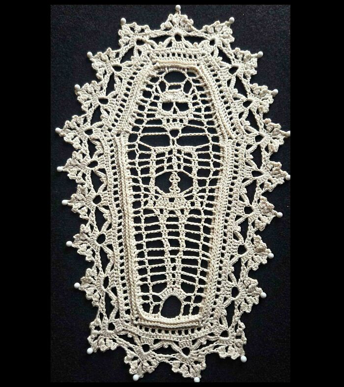 An awesome crocheted coffin-shaped doily in beige, with small white beads adorning the scalloped edges.