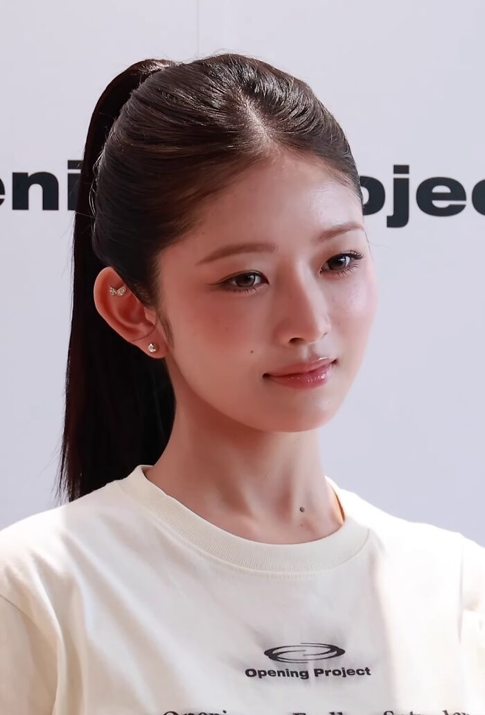 Rei with her hair in a ponytail, wearing a white shirt and small earrings during a public event.