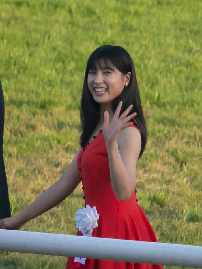 Tao Tsuchiya smiling and waving outdoors, wearing a red dress with a white ribbon at a green grassy event.