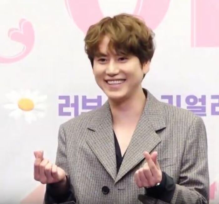 Cho Kyu-hyun smiling and making finger hearts gesture, wearing a gray blazer at a public event.