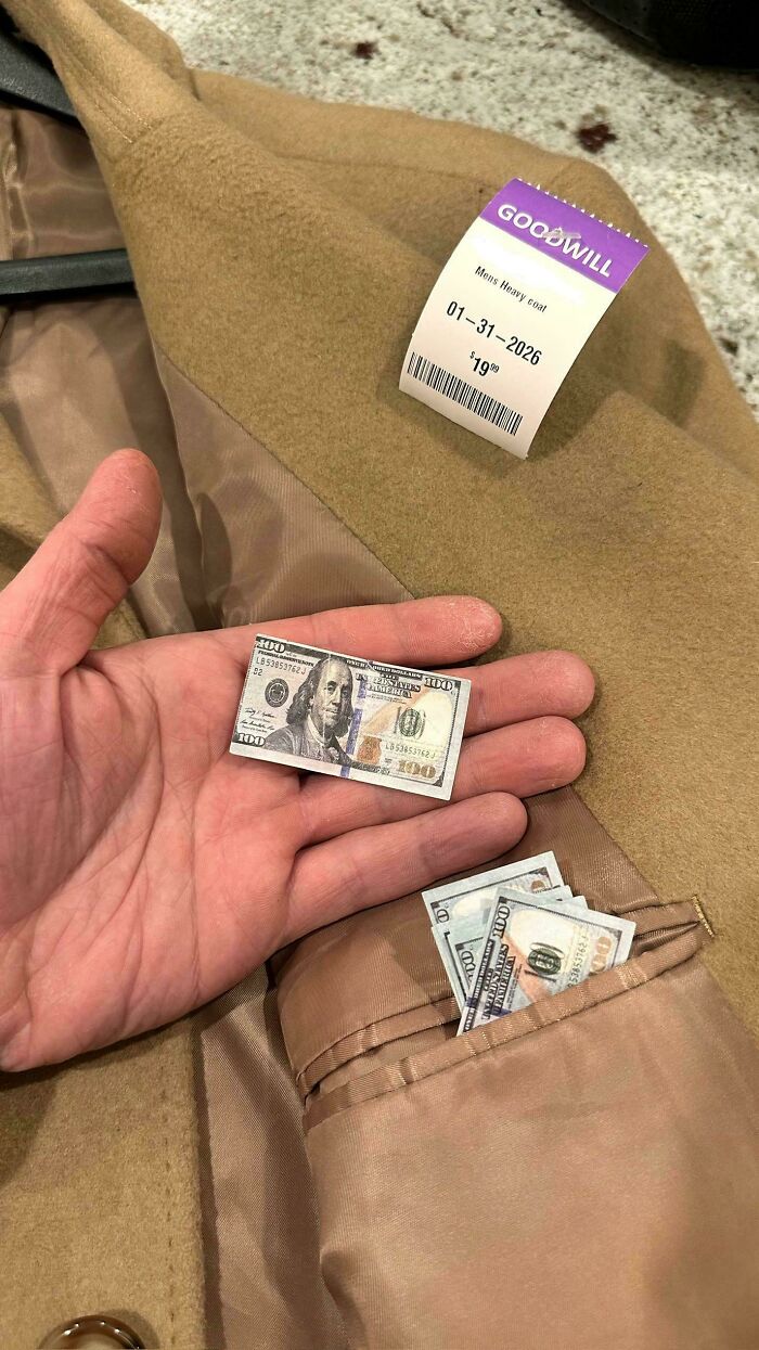 A hand holding a miniature hundred-dollar bill next to a Goodwill coat, a surprising jackpot discovery.