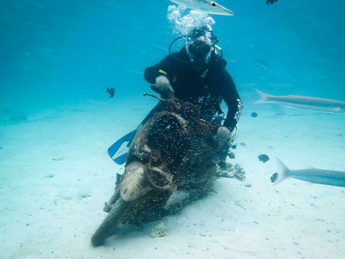 Scuba diver underwater riding an old motorcycle covered in marine growth with fish swimming nearby in ocean photos.