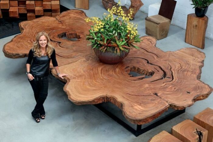 Woman standing beside a large wooden table carved from a natural tree slab by talented woodworkers in a modern workshop setting.