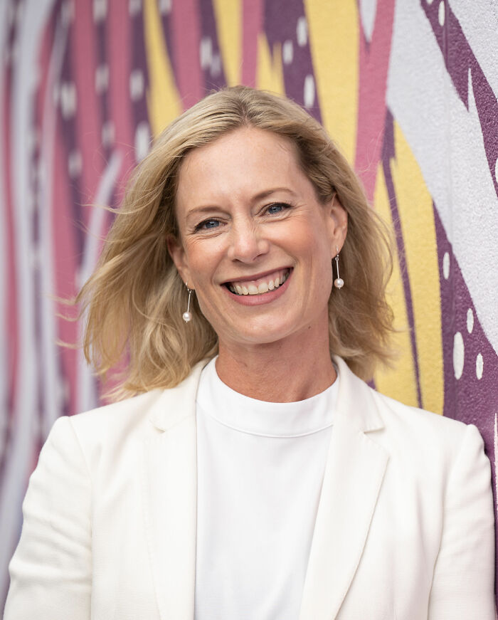 Rebecca White smiling outdoors, wearing a white blazer and pearl earrings, with colorful mural background.