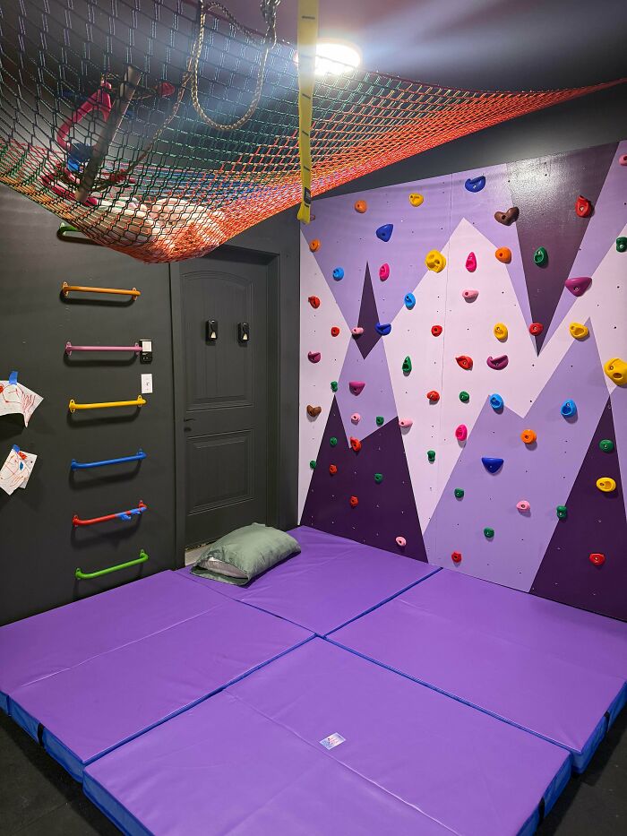 A home climbing wall for kids, featuring purple and grey mountains, a rope net, and soft mats, built by a great dad.