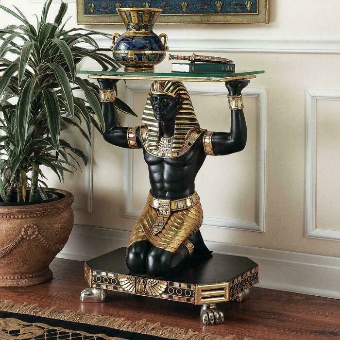 Kneeling Pharaoh statue table holding glass top with decorative vase and plant in a living room with sculpted details.