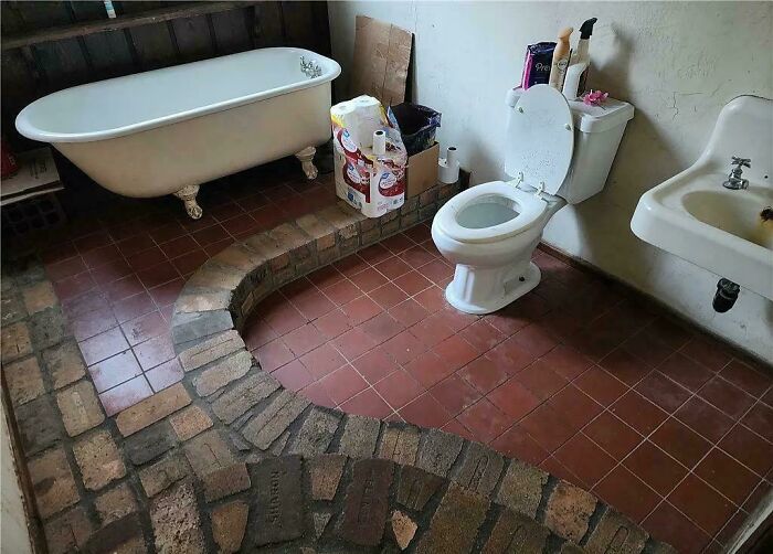 Bathroom with clawfoot tub, toilet, sink, and a peculiar raised brick platform in a rustic setting, showcasing ridiculous home decor.