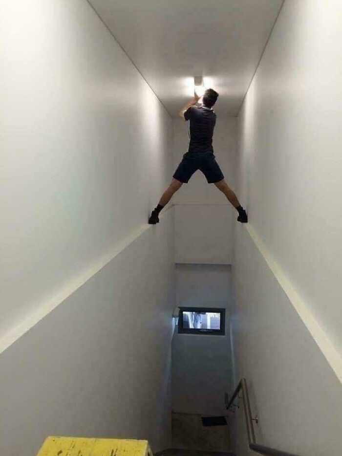 Man precariously balancing on narrow ledges to fix ceiling light in ridiculous home decor setting with sarcastic DIY humor.