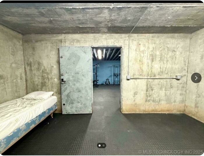 Concrete room with a single bed and an open metal door showing an industrial interior, showcasing ridiculous home decor.