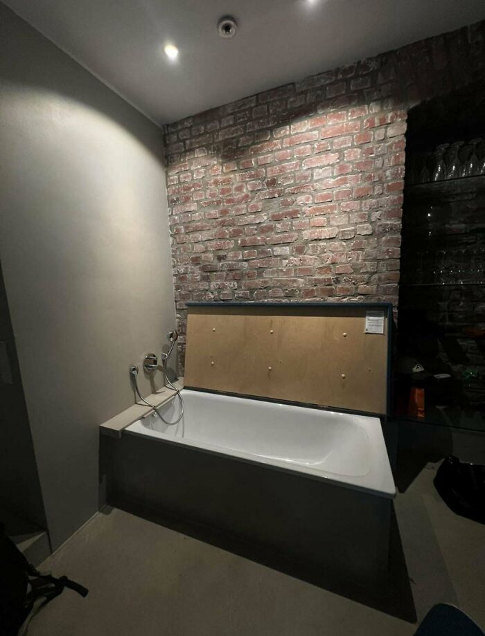 Bathtub with exposed plumbing against unfinished brick wall and plywood panel in ridiculous home decor setting.